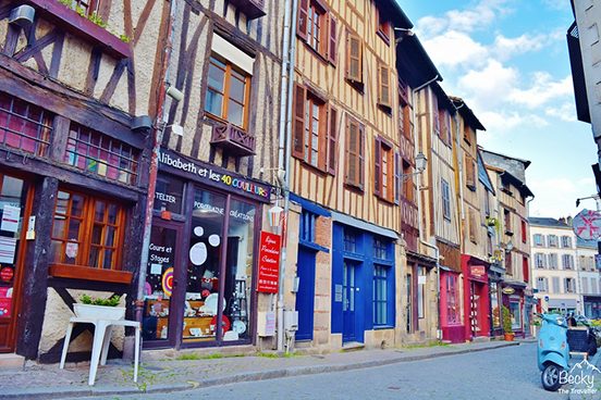 Rue pavée avec maisons à colombages colorées et boutiques, ambiance médiévale ensoleillée.